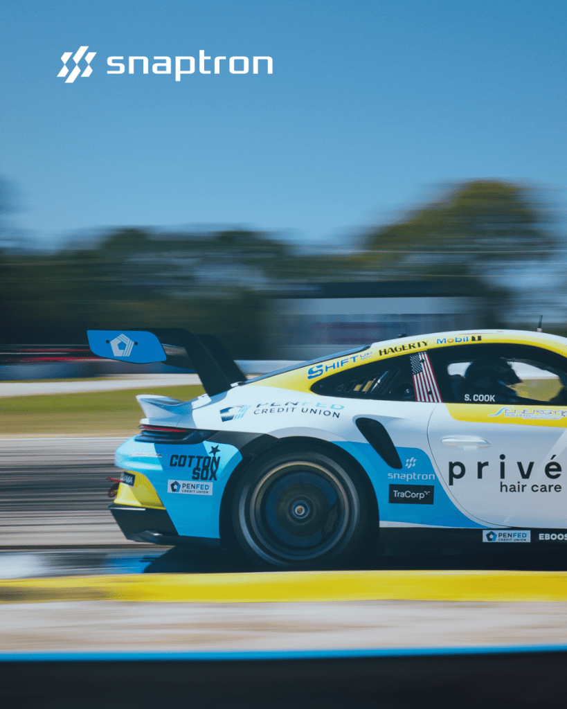A sleek racing car in motion, showcasing the Snaptron logo, sponsors, and vibrant blue and yellow colors, on a racetrack with blurred background.